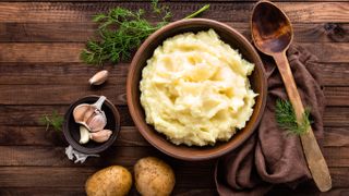 A bowl of mashed potato on a wood surface surrounded by potatoes and garlic