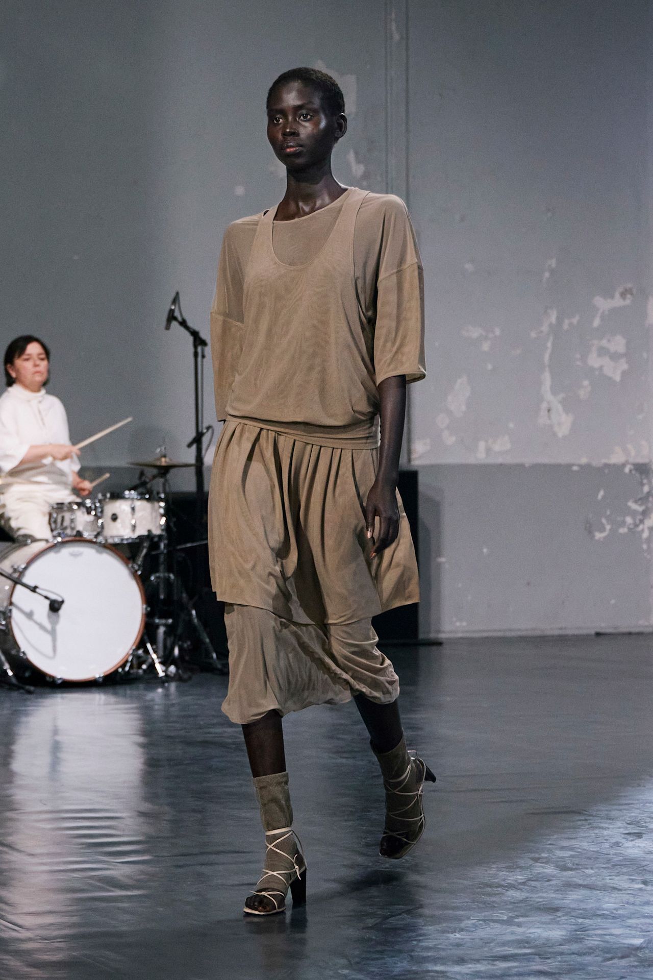 Layering staples for spring 2026 is shown in a photo from Lemaire's runway show, with a model walking while wearing a semi-sheer tan t-shirt layered under a tan tank top styled with a tan skirt, sheer socks, and strappy sandals.