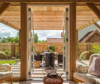 The view out of glass patio doors from a sunny living space. The doors are open and the walls either side are fixed glazing. The patio is partially covered with an open timber pergola which is built in a matching wood to the oak frame of the house