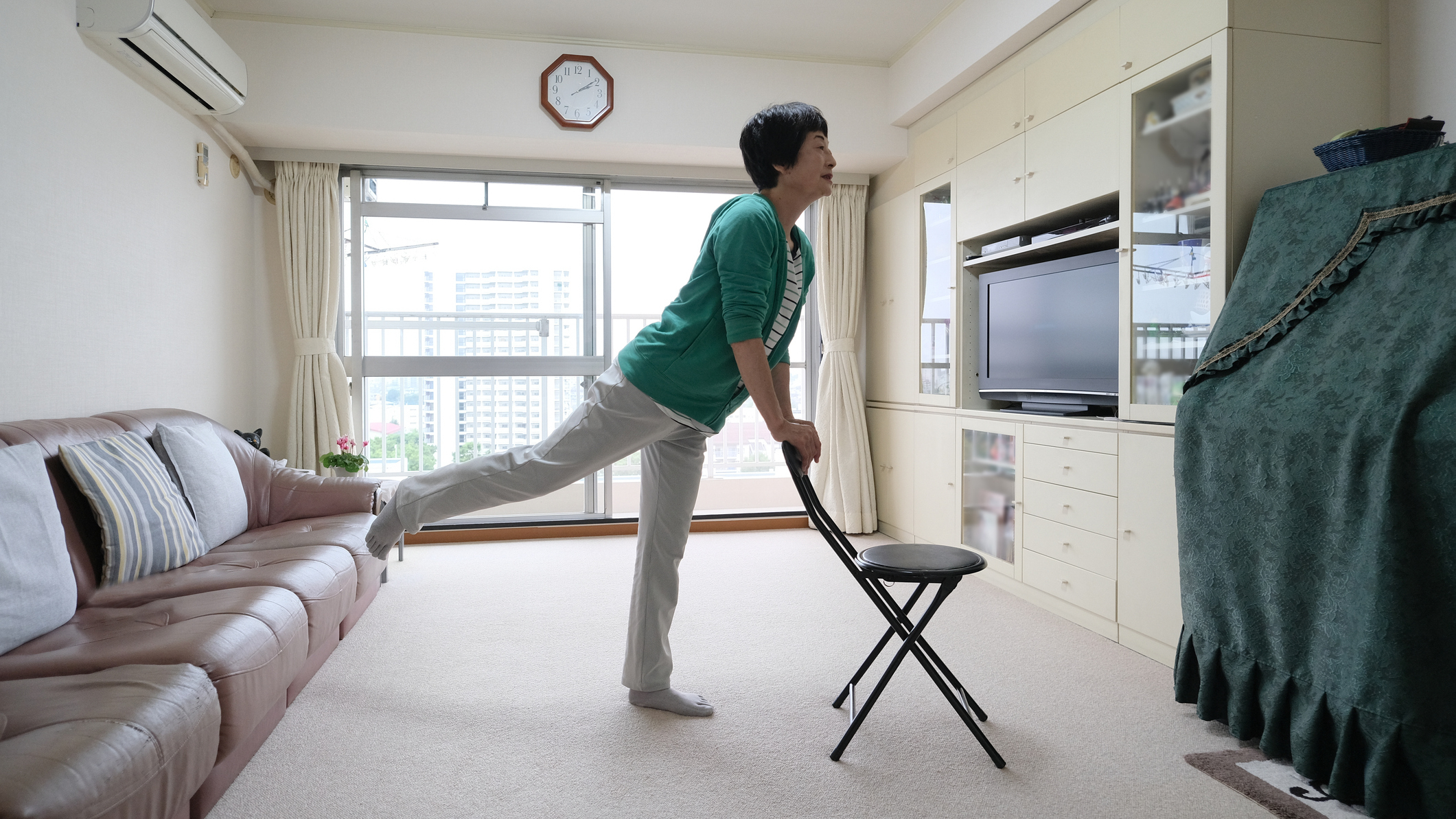 Senior woman exercising in living room holding back of a chair and lifting one leg behind her
