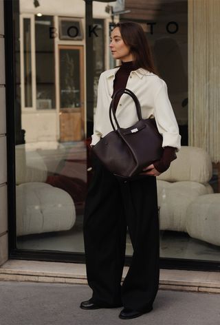 A cute work winter outfit is shown in a photo of a woman standing on the sidewalk in Paris wearing a dark red turtleneck sweater under a white button-down shirt with black trousers, loafers, and a dark red handbag