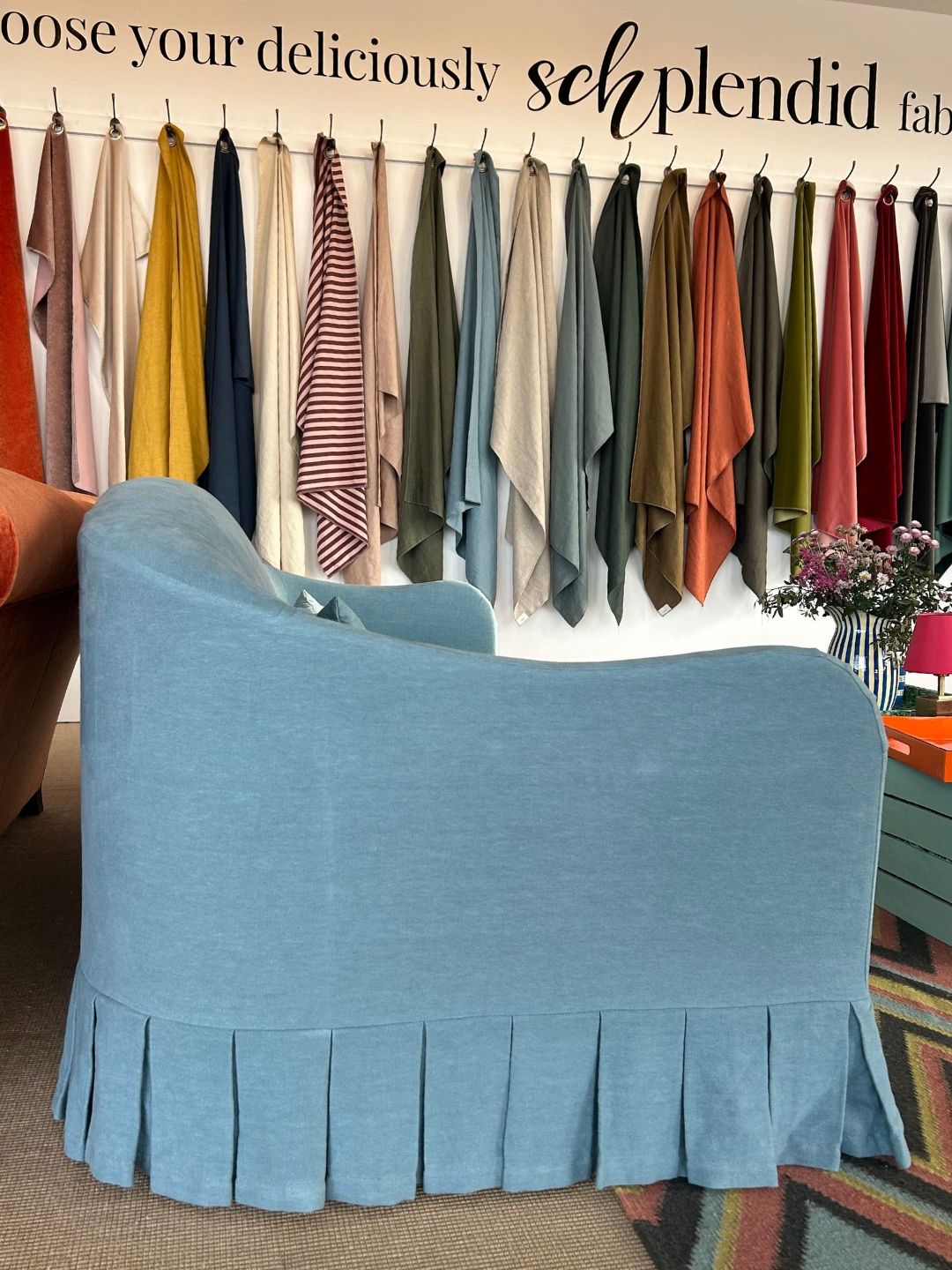 Side view of Schplendid's Edie sofa in pale blue Skye linen, showing its curved camelback, rounded silhouette and box-pleated skirt, with fabric samples displayed behind.
