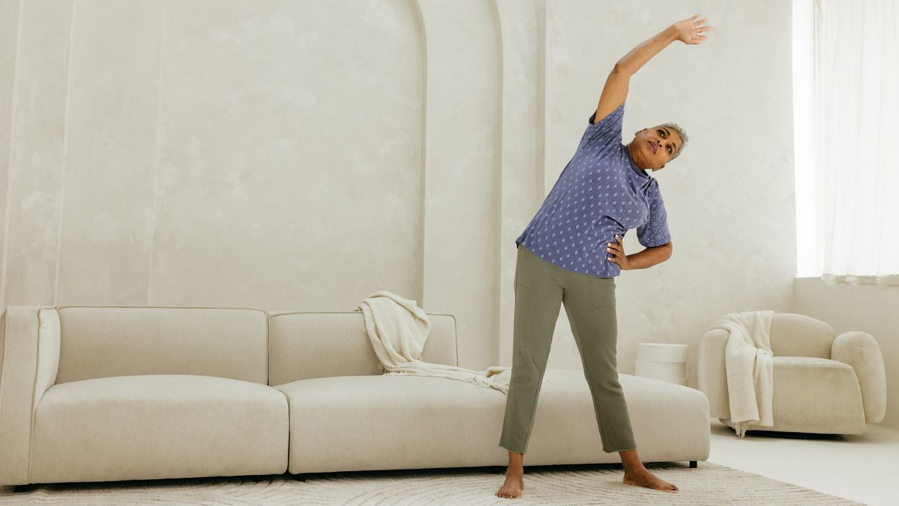 Senior woman reaches her right arm above her and to the left. She stands in front of a white couch 