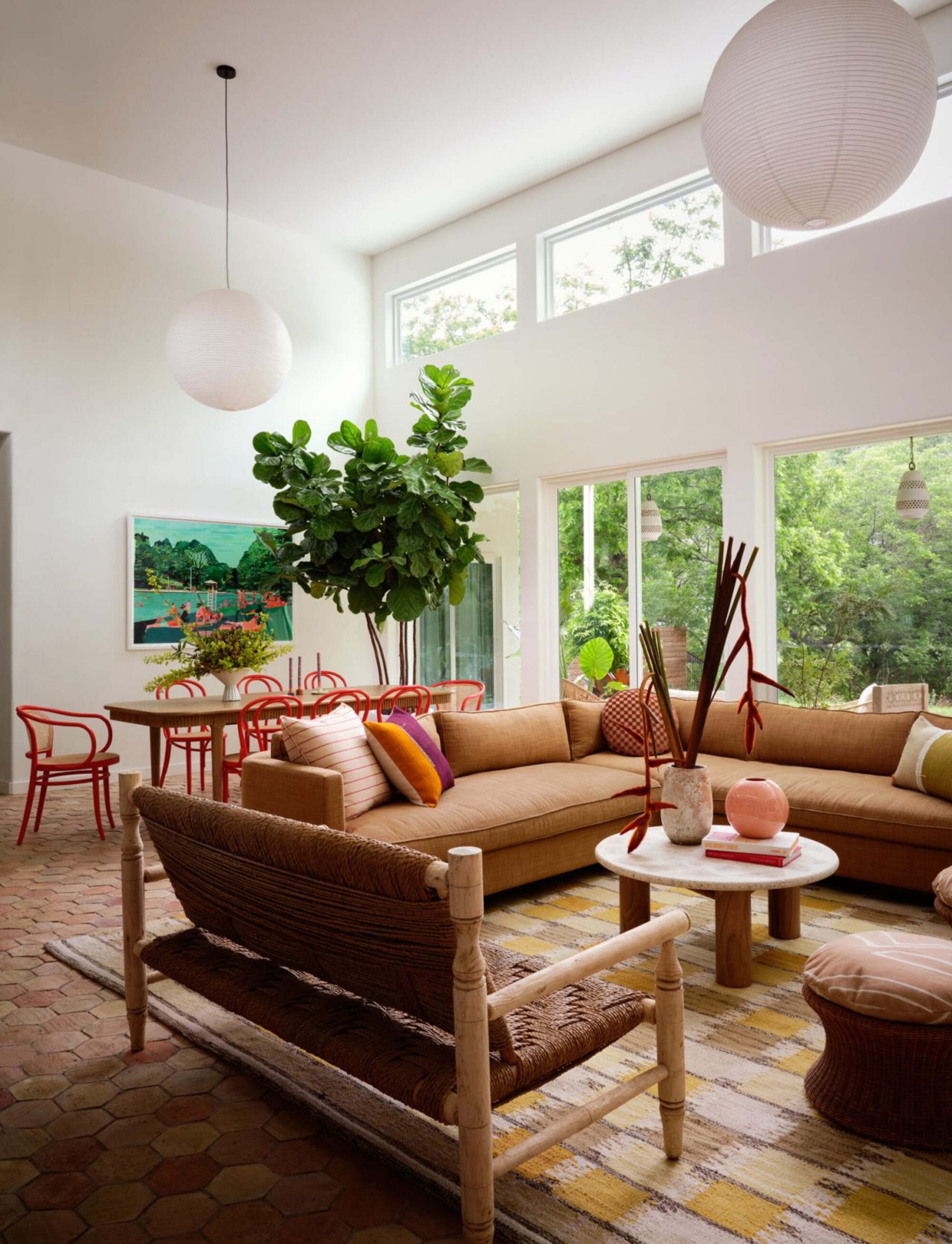 colorful graphic rug in open-plan living room with tan L-shaped sofa, leather and timber benchseat, and round coffee table styled with vases, with a timber dining table in the background with red dining chairs, paper pendant lights hanging, terracotta floor tiles, and large windows out to the garden