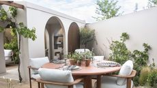 A round outdoor dining table with four chairs on a modern patio with white walls and archways