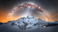 Snowy mountains under a vibrant, starry sky with the Milky Way arching overhead. A glowing tent sits to the right, creating a sense of solitude and wonder