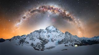 Snowy mountains under a vibrant, starry sky with the Milky Way arching overhead. A glowing tent sits to the right, creating a sense of solitude and wonder