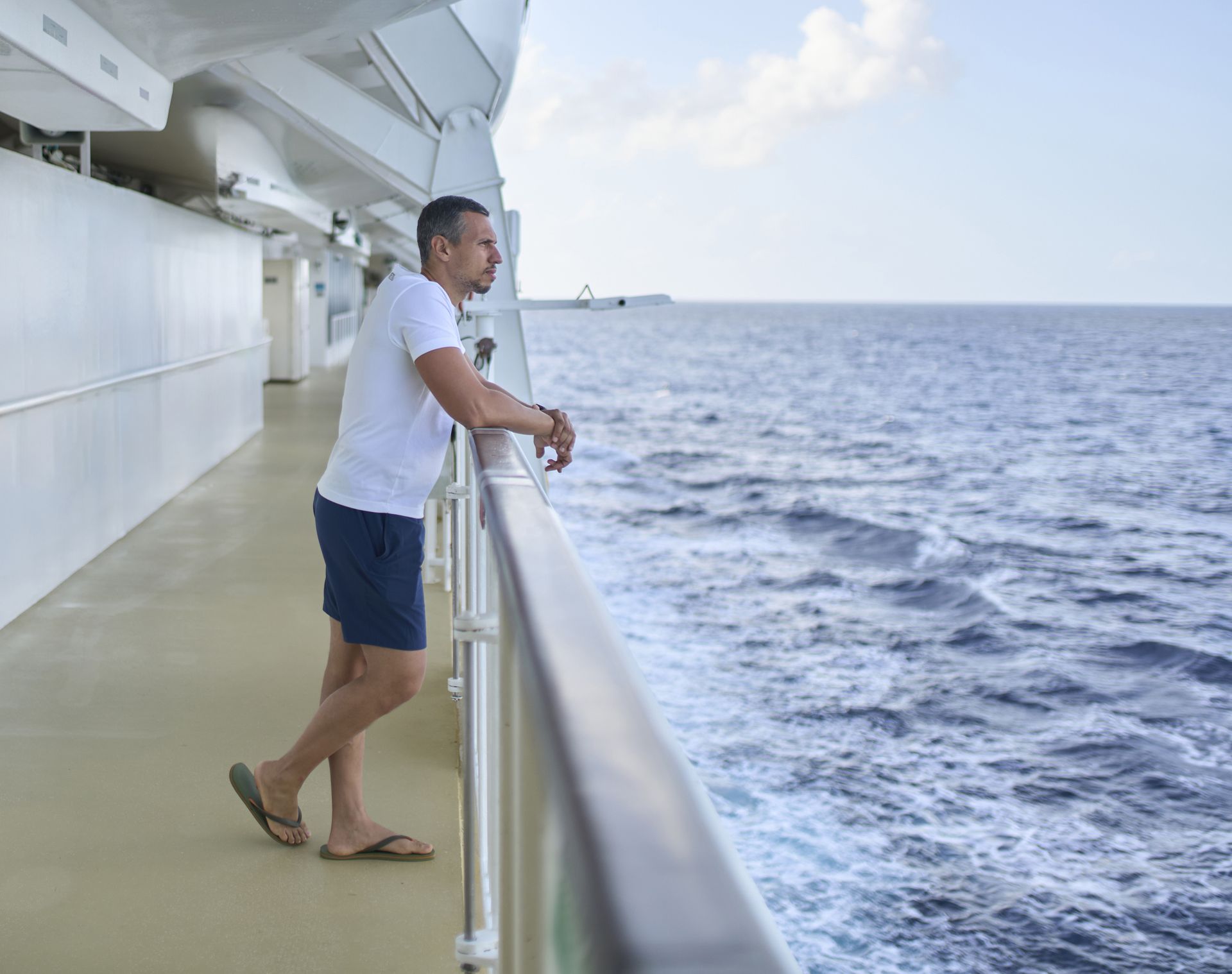 Side view of a man in a white t-shirt standing at the railing of a cruise ship deck, gazing at the ocean horizon.