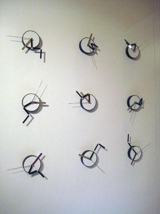 Nine mini black and white clocks hanging on a white wall.
