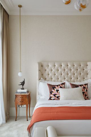 beige bedroom with upholstered bedhead, bold print retro pillows, antique side table, pendant light, rust blanket