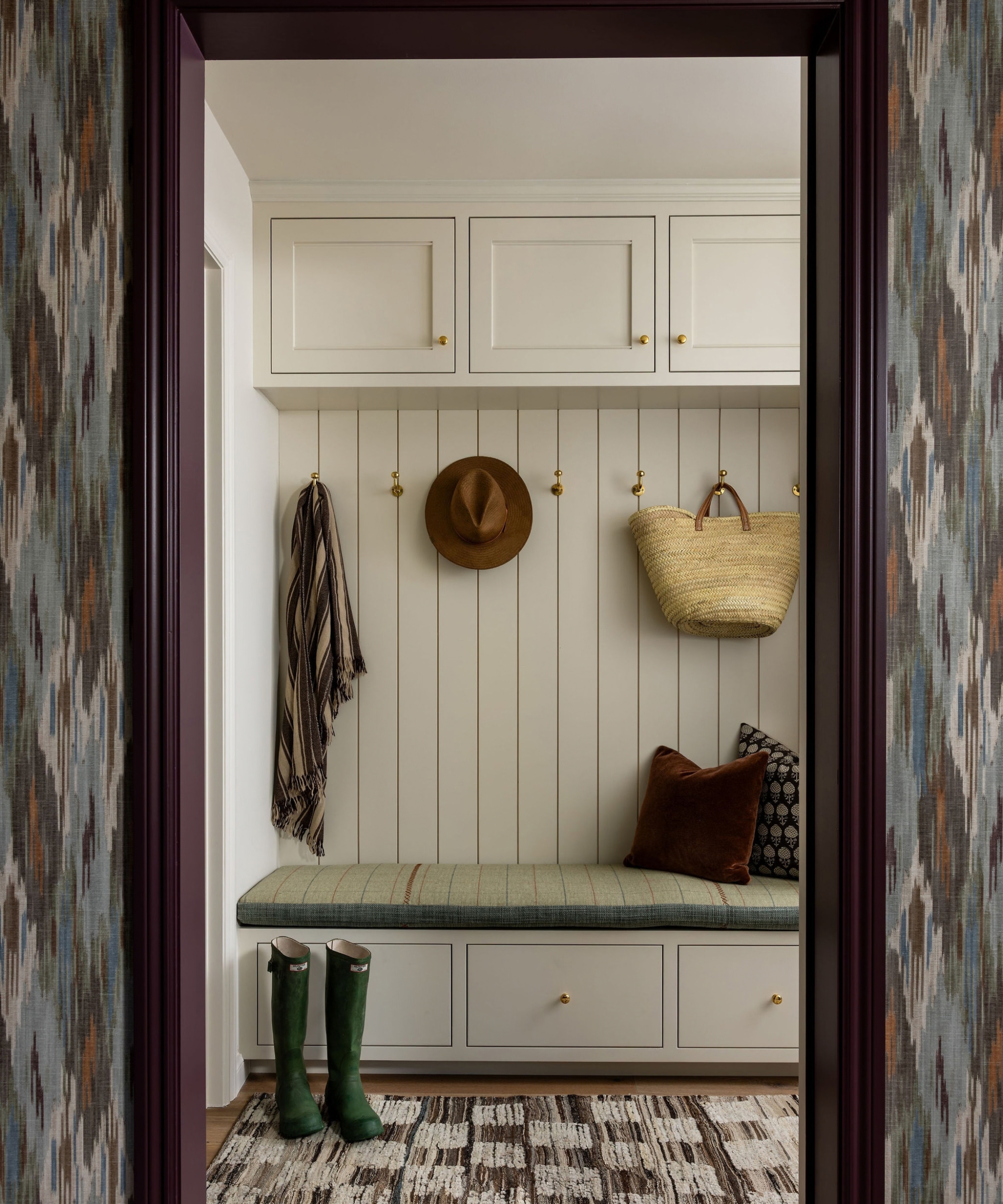 Mudroom with cream cabinets, brass hooks and a cushioned bench, viewed through a plum doorway framed by patterned wallpaper.