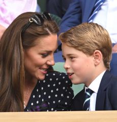 Prince George wearing a blue suit leaning over and talking to mom Kate Middleton, wearing a blue polka dot dress, at Wimbledon 2022