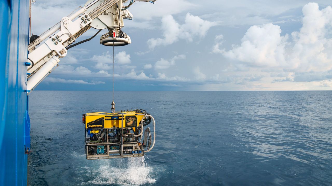 Underwater vehicle being hoisted out of sea by crane aboard boat.