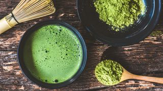Matcha tea powder sitting in a bowl next to powder combined with water with whisk on dark wooden coffee table