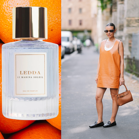A woman next to a bottle of perfume wearing an orange dress and black loafers is holding a brown handbag. She is also wearing sunglasses as well as a slicked-back bun. 
