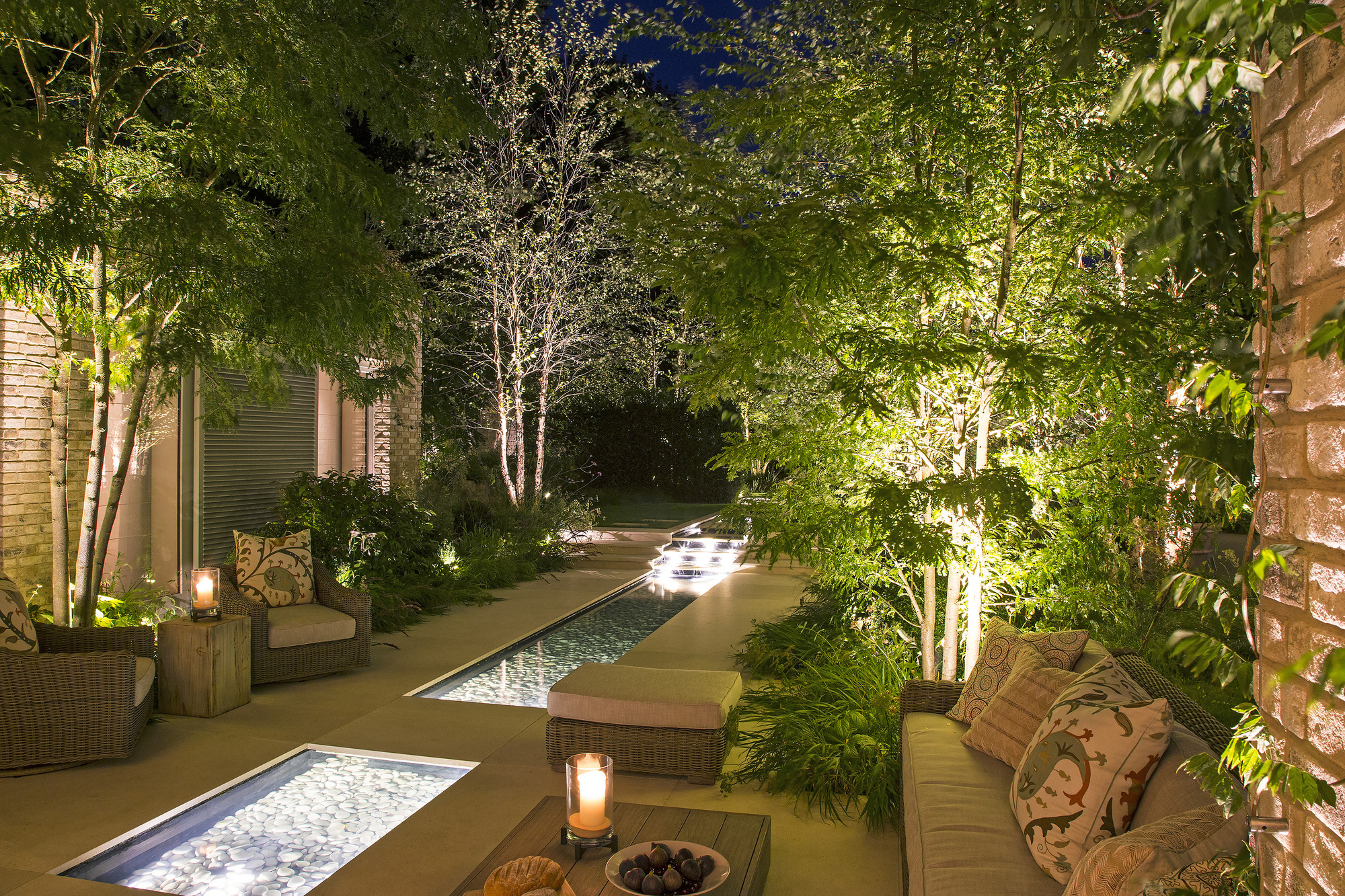 A modern garden at dusk with water feature pools that are lit up, and trees with uplighting lining the walls