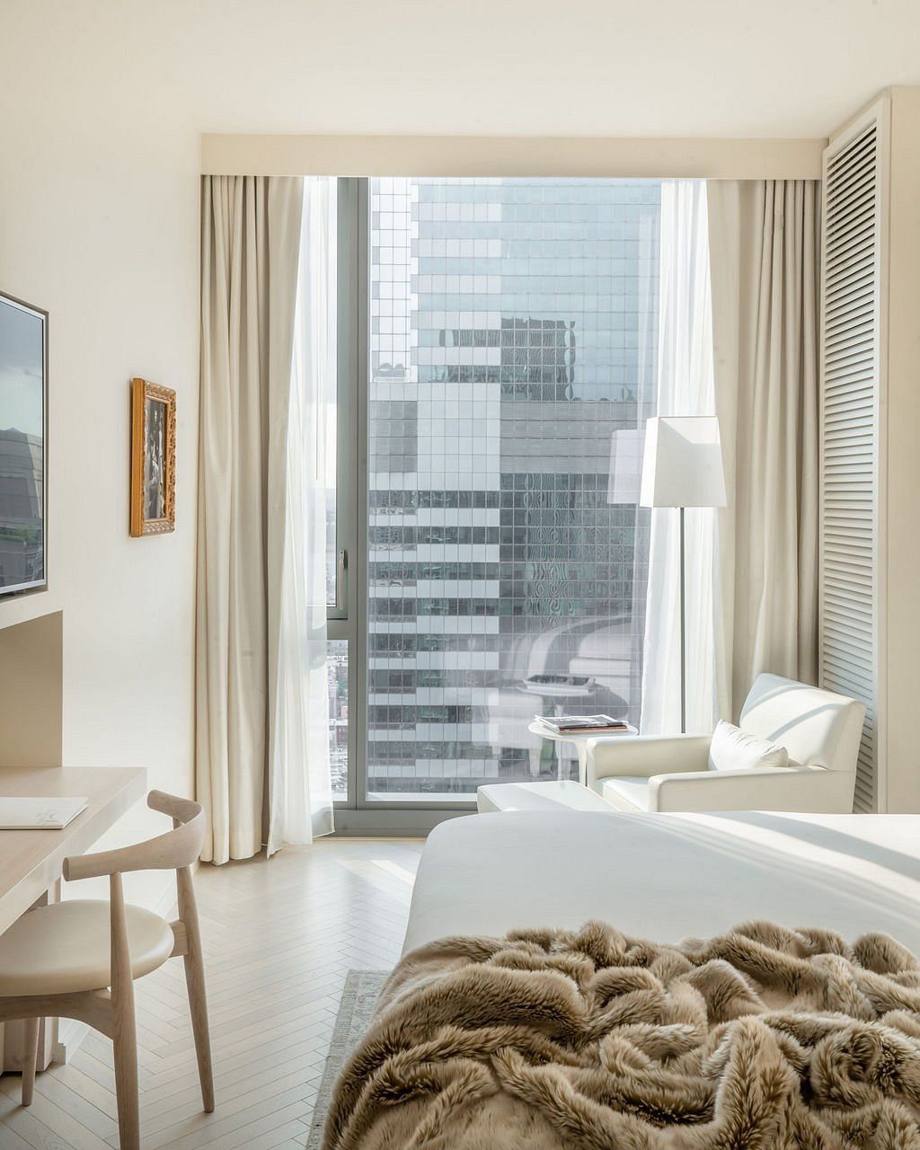 A superbly decorated, hyper-modern hotel features minimalist cozy furniture including neutrally-hued large couches, standing lamps, tiled floors, cream curtains, wall-to-wall windows, and breathtaking views of the city.