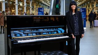 Claudia Winkleman stood next to an upright piano in a train station 