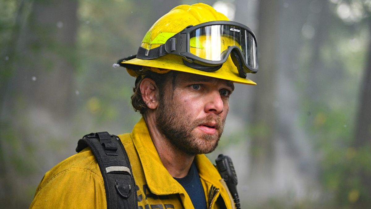 Max Thieriot as Bode standing among the trees wearing a yellow hardhat and jacket. He looks serious.