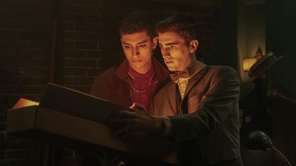 A still image of Charles and Edwin from Dead Boy Detectives, in dark room, looking in a book that is somehow emitting a glow of light