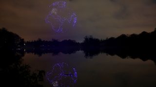 A series of purple drones come together in the night sky above a still lake to create a 3D image of planet Earth with the continent of Africa seen and reflected in the lake