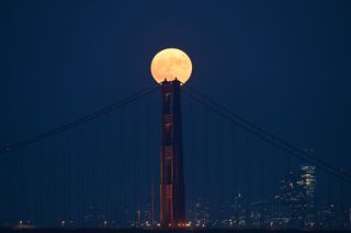 bright full moon shines above the center column of the golden gate bridge, with the city skyline in the background.