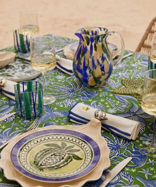 An outdoor dining table with a green and blue tablecloth, a blue and green pitcher and wine glasses