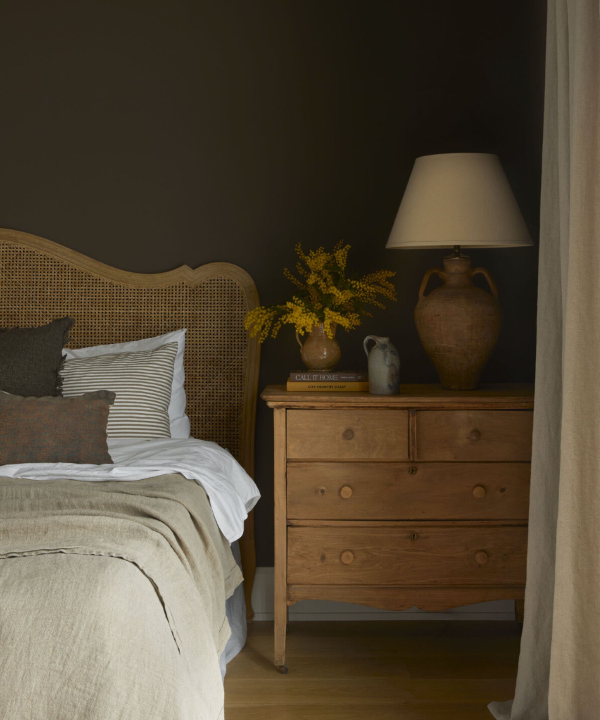 Cozy brown bedroom with rattan headboard, white and cream bedding, brown and cream pillows. Wooden cabinet next to bed with books, lamp, vase of flowers, warm wood flooring