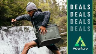 Fisherman striding across rocks by waterfall carrying Yeti soft cooler in Camp Green