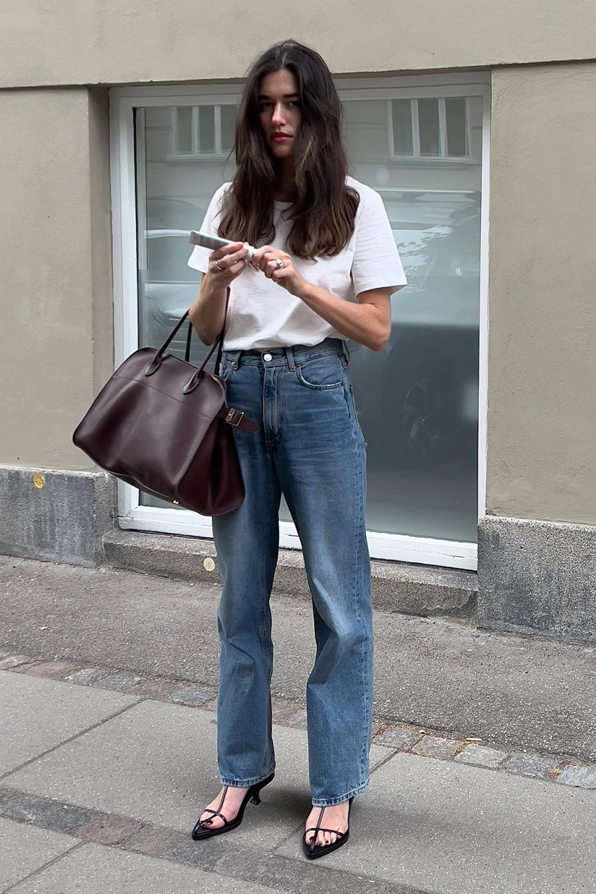 A woman standing on a sidewalk wearing a white t-shirt tucked into a pair of baggy jeans styled with a pair of strappy sandals and a brown bowler bag.