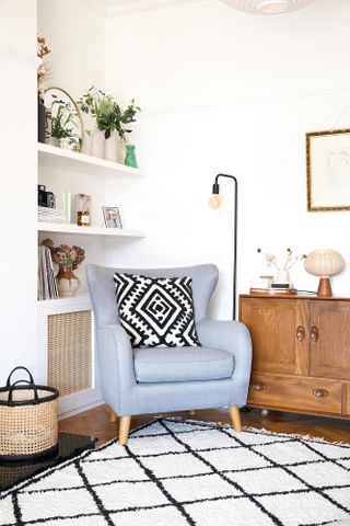 Corner of living room with white walls, black and white rug, light blue armchair, black floor lamp and white shelves filled with accessories and magazines