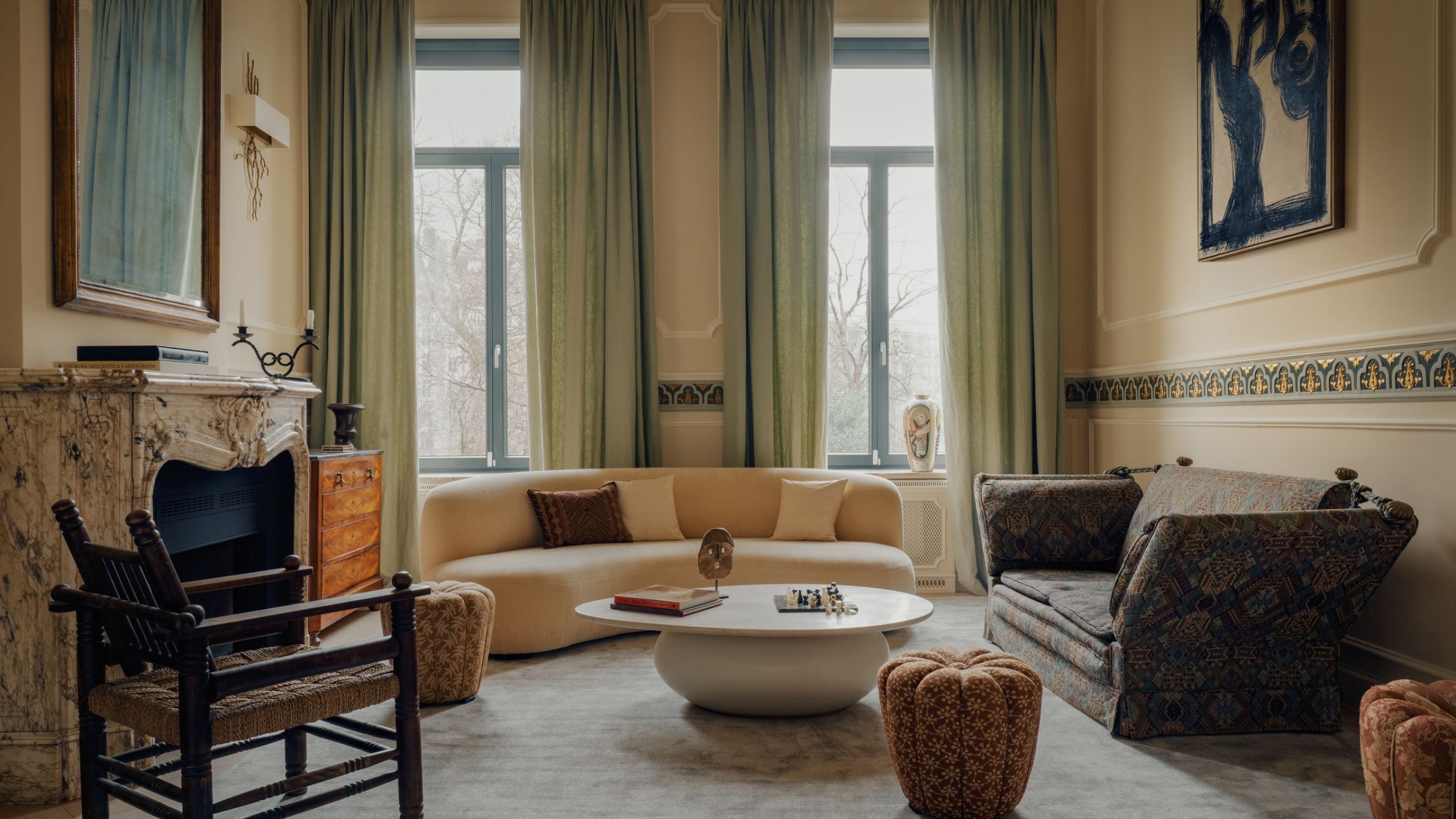 modern living room with sage green curtains, yellow walls, marble fireplace, curbed modern sofa, brown sofa, round coffee table, ottoman, and timber-and-woven armchair