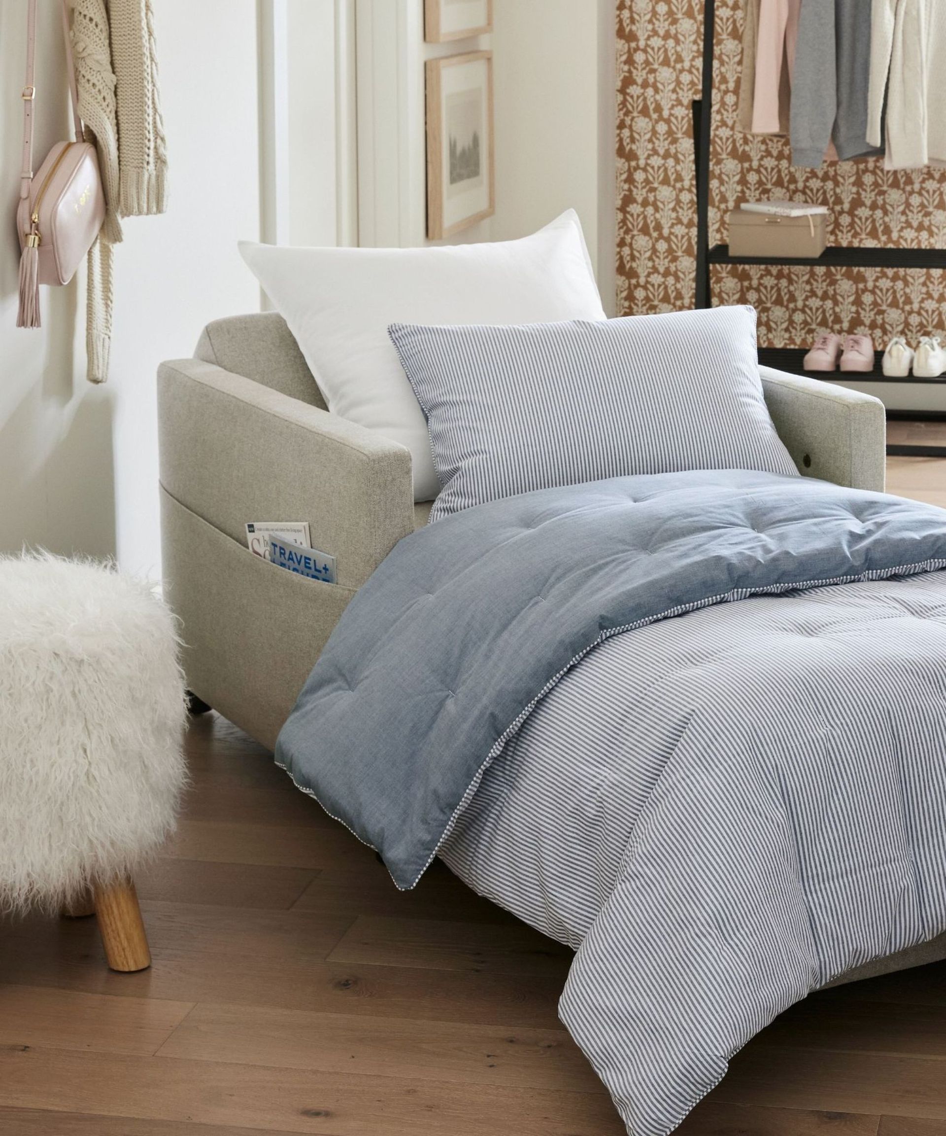 pottery barn light grey armchair that folds out into a sleeper sofa bed styled with blue and white striped bedding with a sheep skin sherpa footstool sat next to it