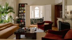 Image of a large magazine rack leaning against the wall of a grand living room. There is a sitting area with a long, brown leather sofa, a black marble coffee table, and two red reading chairs.