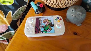 Anbernic RG Cube on pine bedside table next to Frog jewellery pot and Pingu ornament with green bedsheets on left.