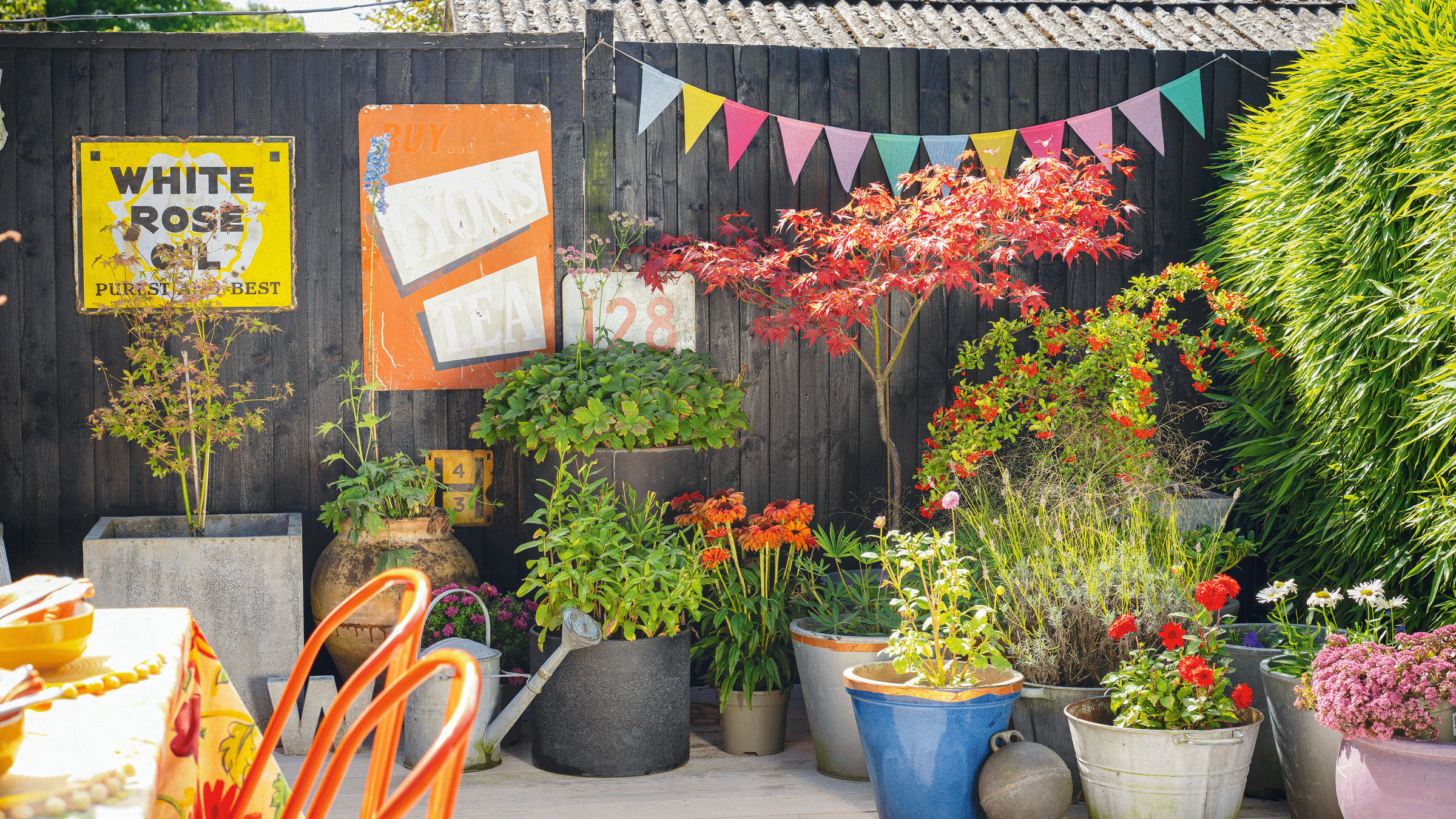 a patio with a black painted fence with large vintage signs and bunting with lots of planters filled with plants and flowers beside a dining table and chairs