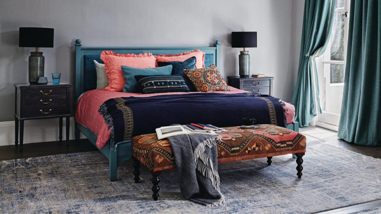 Showing off the best bedframe: a bedroom with grey walls, teal blue curtains and grey rugs on a wooden floor, colourful red and blue bedding and brown chest of drawers.