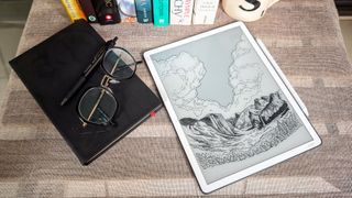 A wallpaper displayed on the 3rd generation Kindle Scribe lying on a table beside a notebook, glasses and some books