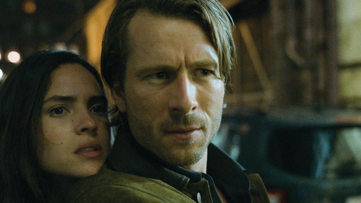 Glen Powell looking concerned with his co-star Adrina Arjona standing behind him in Hit Man.