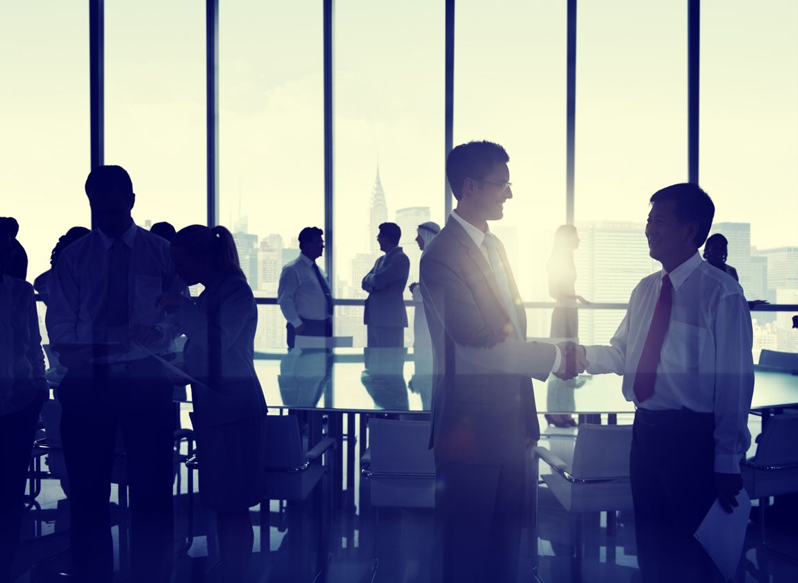 Business people shaking hands in an office building against a New York skyline.There is a large desk in the center of the room surrounded by office chairs.