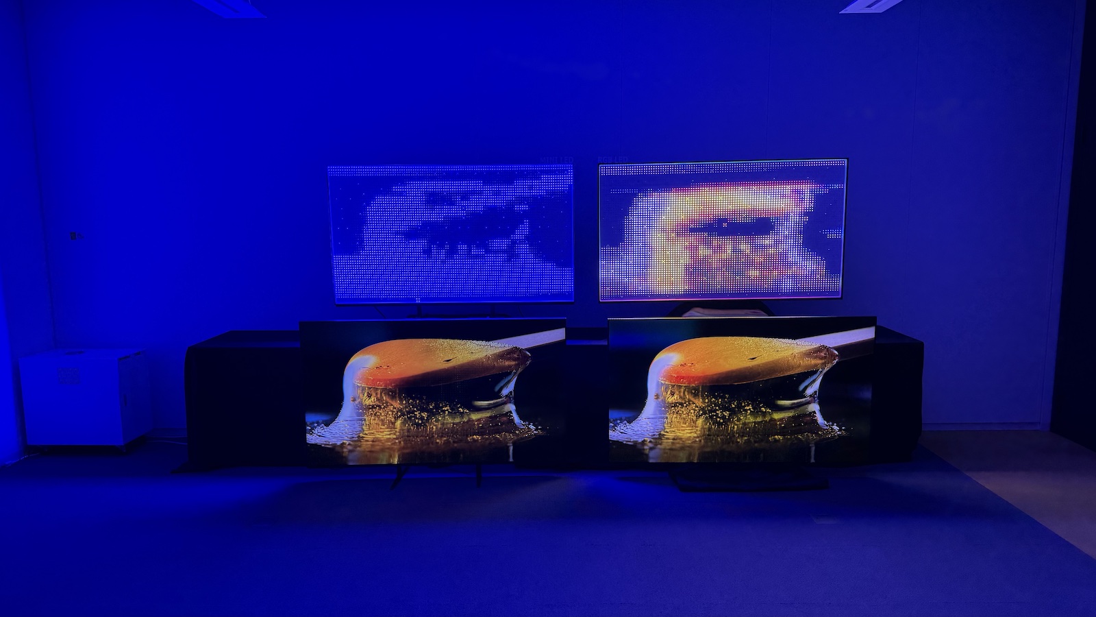 Two TVs arranged next to one another on the floor, with the exposed backlights of each on a table above. On both of the assembled TVs, an image of a thick golden liquid is displayed. On the left-side backlight, a vague blue image is visible. On the right-side backlight, the colours are fully recreated and the shapes of the image are clear.