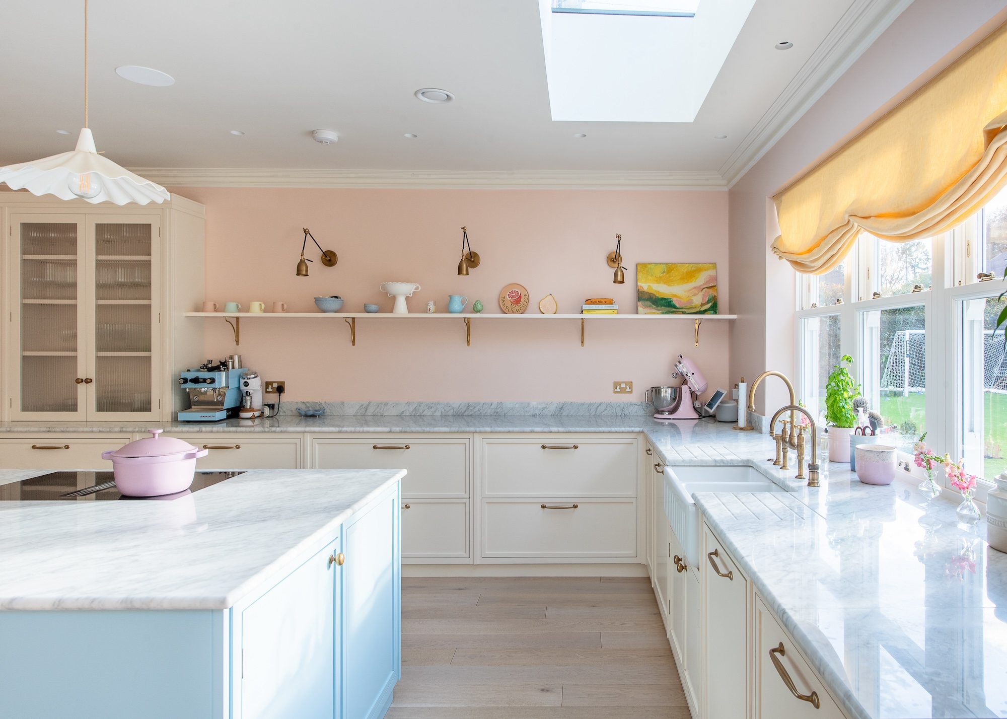 A light kitchen with pale pink walls and glossy marble work tops, there is an island to the left wit a window on the right above the sink, and cream and power blue cupboards.
