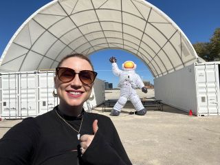 a woman in sunglasses smiles in front of a series of white shipping containers and a large inflatable astronaut in a white spacesuit