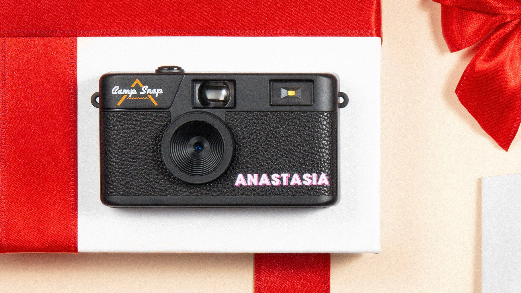 A lifestyle image of the Camp Snap Custom camera with personalized name 'Anastasia' on the front of it in block capitals and a pink color. It's a Christmas themed product image with a red present and bow background design