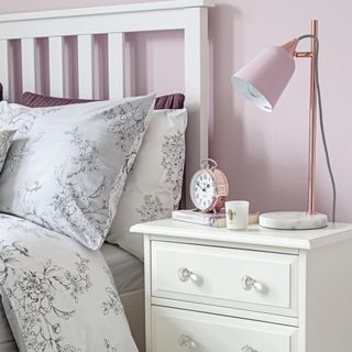 A chest of drawers with glass knobs, cushions and table lamp in a cosy blush pink bedroom corner.