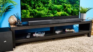 The Polk Audio Signa S4 being used in a lounge with a large TV. On the TV screen is a green forest scene. In the background is a blue wall.