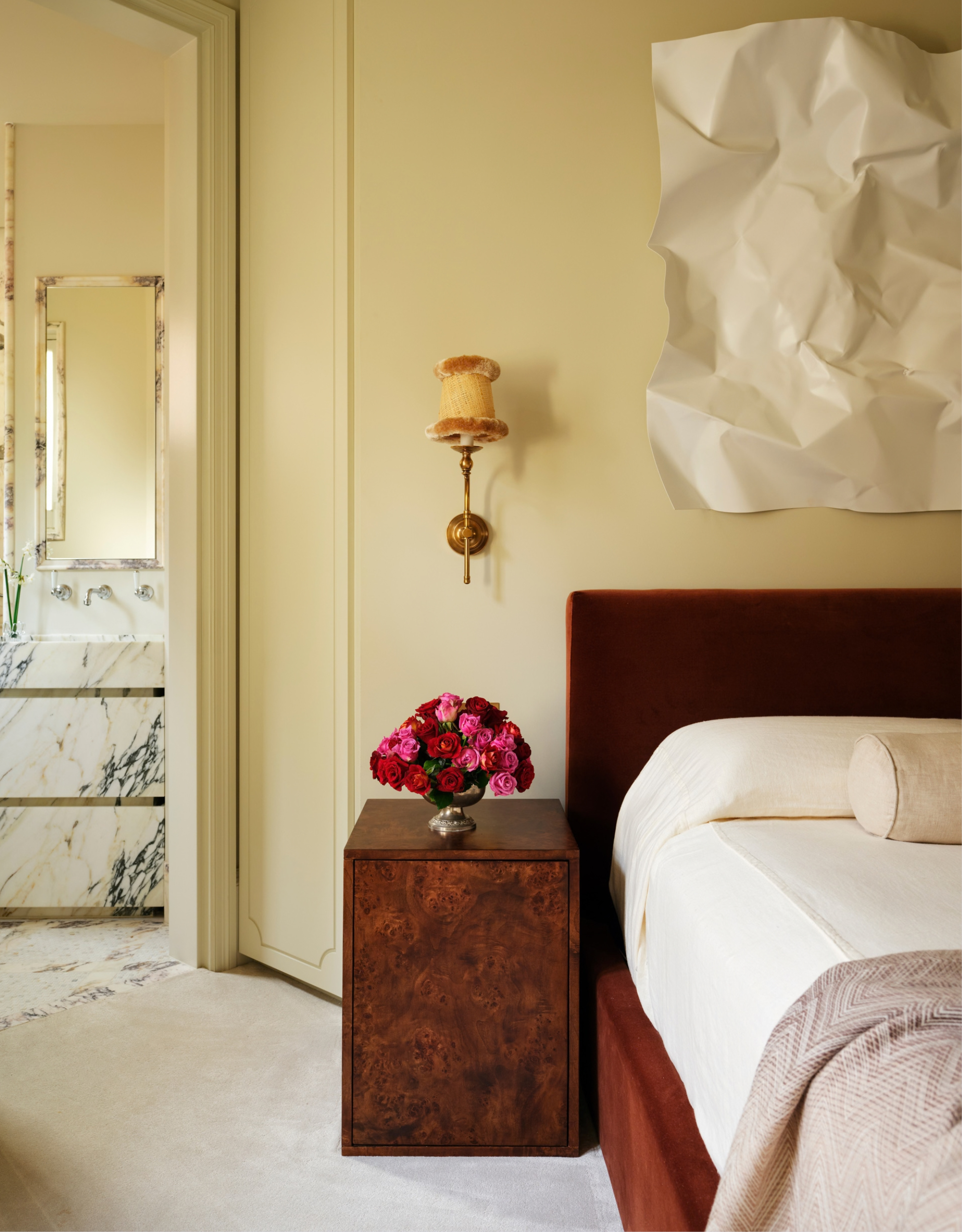 A beautiful bedroom with a textured white art piece on the wall, a tufted rim wall sconce, a burl wood nightstand, a white bedscape, and a marble bathroom
