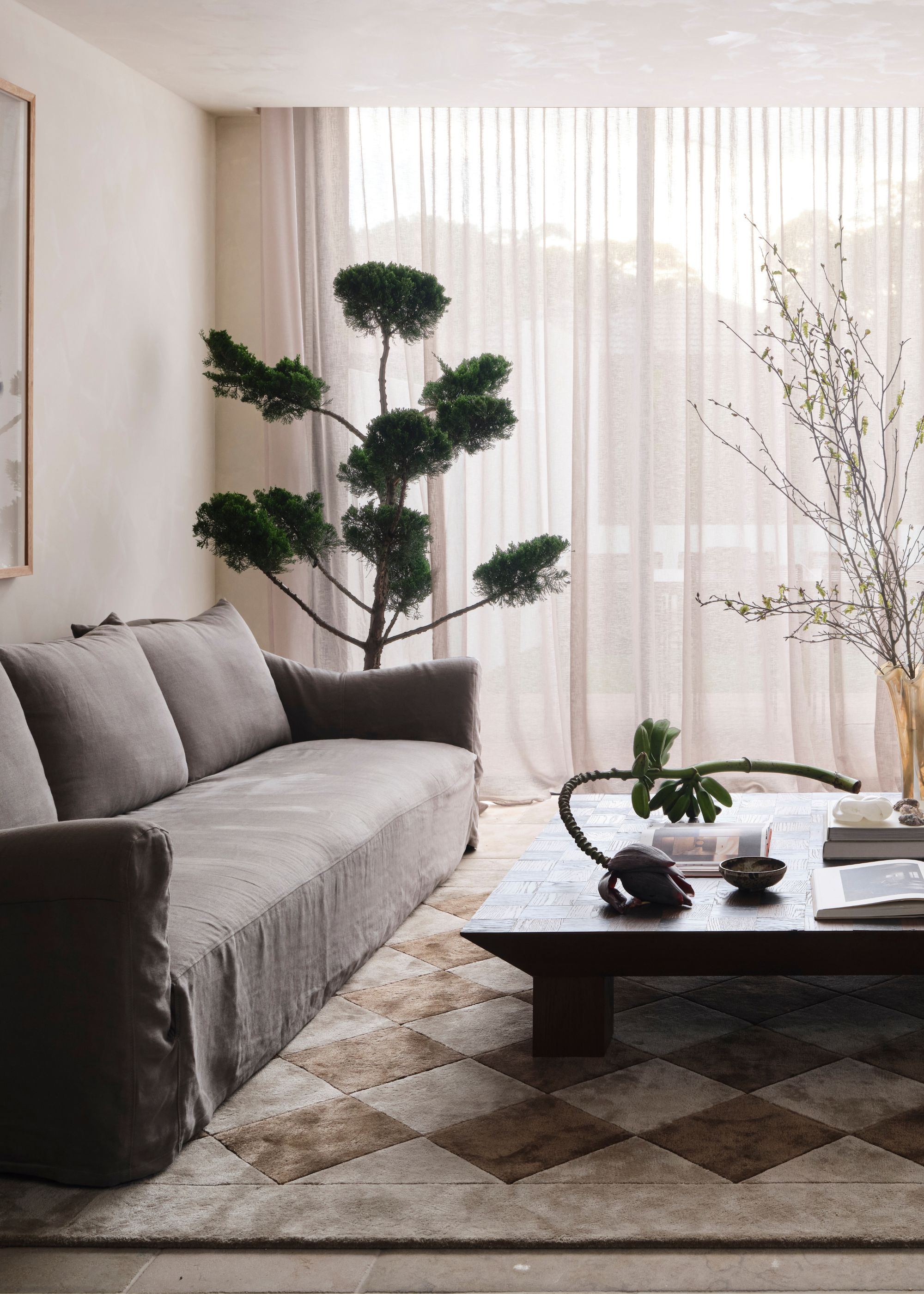 A muted living room with a potted tree, a vase of stems, a plantain branch, a low coffee table, a gray couch, and a checkerboard rug