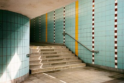 Stairs in the subway of a train station featuring, grey floors and step, silver steel banister and green tiled walls 