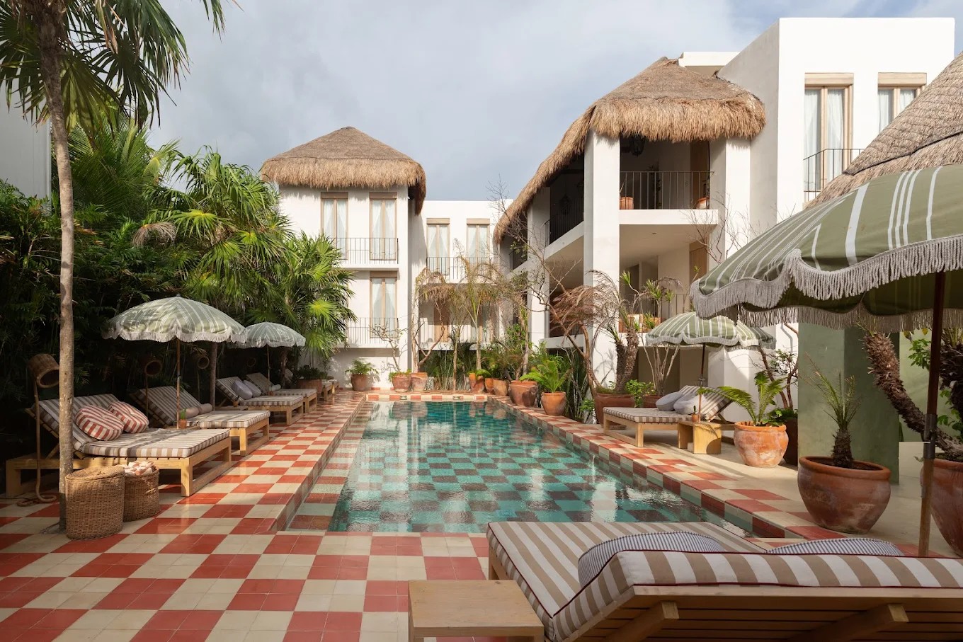 A checkered pool in pale red and white tiles, surrounded by vintage green and white parasols and tropical architecture in white cement with decorative palms.